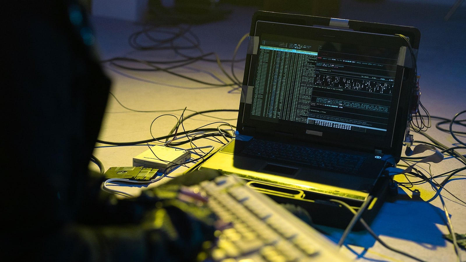 Notebook com telas de terminal e código em um ambiente escuro, cercado de cabos